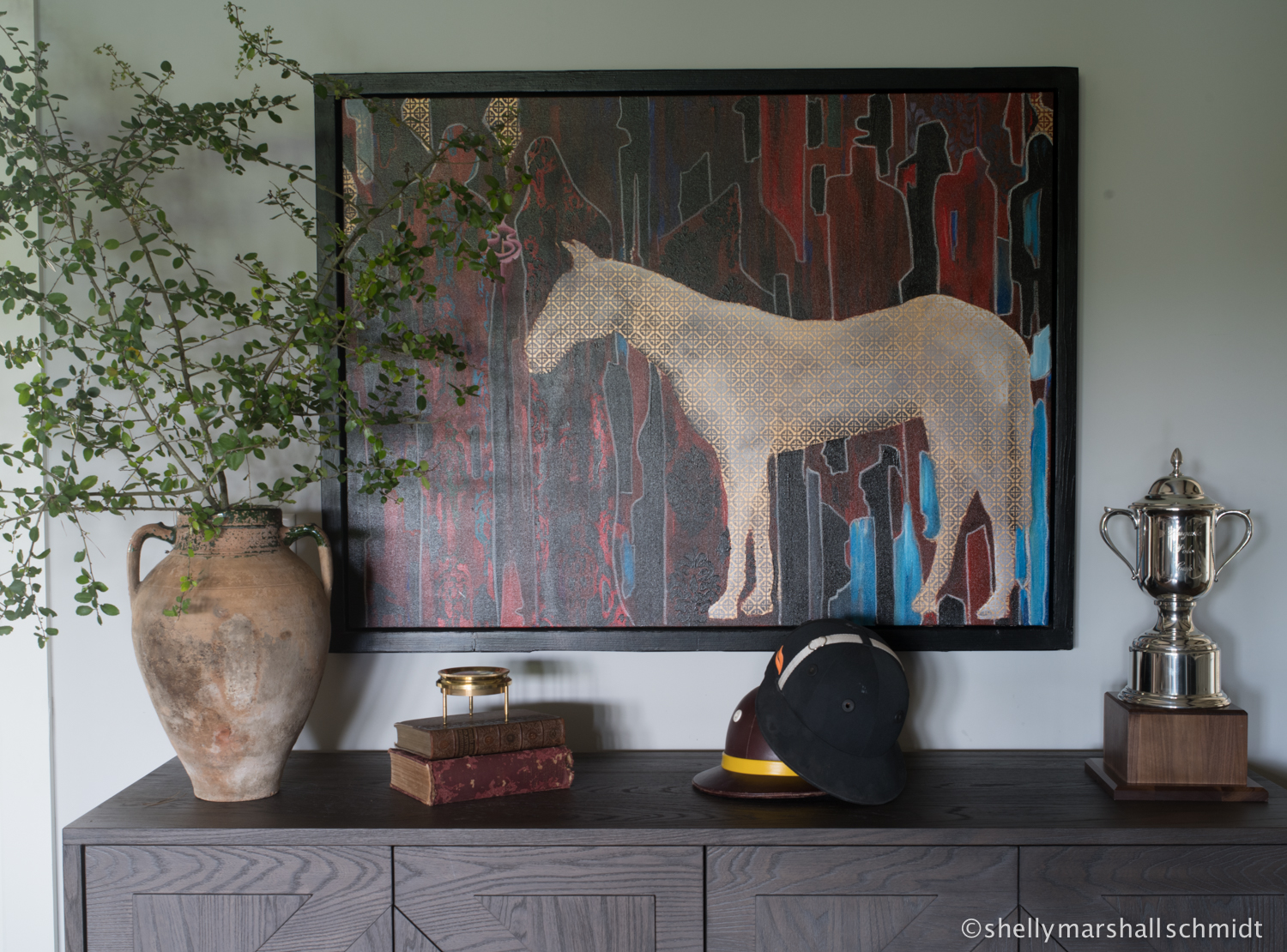 Console table styled with equestrian artwork, vintage trophies, a riding helmet, books, and an oversized urn with greenery.